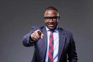 confident african businessman in suit and tie pointing finger at viewer on clear grey background photo