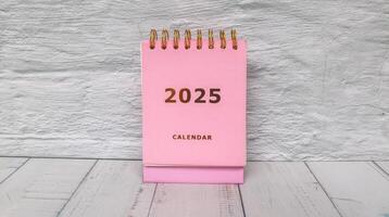 Pink calendar 2025 standing on office desk with flower pot and alarm clock. New year 2025 with office desk concept with table background. photo