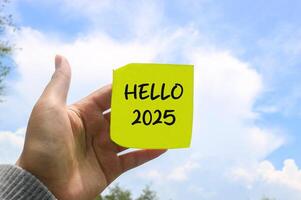 Hand holding a note that says Hello 2025 with a sky background. New year concept with typographic 2025. Motivational words for the new year 2025 photo