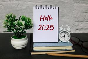Notebook standing on a book with the words Hello 2025 written on it is surrounded by books, alarm clock and flowers on the table. New Year 2025 concept, welcoming the new year 2025 photo