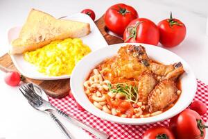 Fresh Tomato Soup with Pork Chop, Chicken Wings, Penne served in bowl alongside scrambled eggs and toast isolated on napkin side view of hong kong food on white background photo