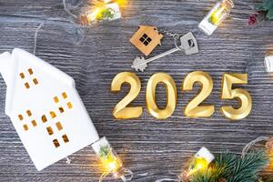 New Year 2025 golden numbers and House key with tiny form of home on festive grey wooden background flatlay, lights of garlands, greeting card. Housing, real estate, relocation, mortgage, insurance photo