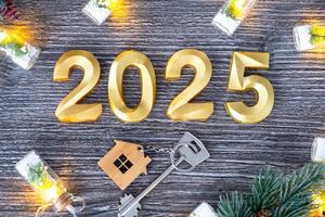 New Year 2025 golden numbers and House key with tiny form of home on festive grey wooden background flatlay, lights of garlands, greeting card. Housing, real estate, relocation, mortgage, insurance photo