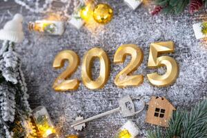 New Year 2025 golden numbers and House key with tiny form of home on festive grey wooden background flatlay, lights of garlands, greeting card. Housing, real estate, relocation, mortgage, insurance photo