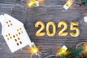 New Year 2025 golden numbers and House key with tiny form of home on festive grey wooden background flatlay, lights of garlands, greeting card. Housing, real estate, relocation, mortgage, insurance photo