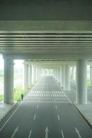 A long, straight road passing through a concrete overpass structure with multiple arched openings, surrounded by green foliage at new yogyakarta international airport photo