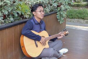 Asian man plays guitar in a park on a sunny day, surrounded by trees. The calm atmosphere and simple beauty of the moment make for a peaceful scene photo