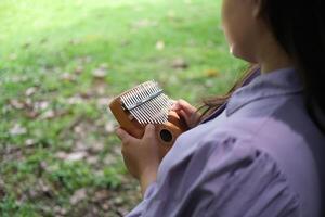young Asian woman plays a kalimba while relaxing in a green city park during summer. The bright sunlight, lush surroundings, and soft melodies create a calm and refreshing outdoor scene photo