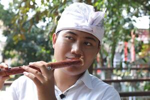 Portrait of Asian man wearing balinese traditional cloth playing bamboo flute in outdoor park photo