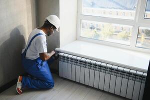 afro repairman in blue using tools while installing or repairing heating radiator. plumber installing heating system in apartment. photo