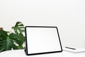 A neat office setup with a blank tablet screen, green plant, notebook and paperwork on a textured white desk. photo