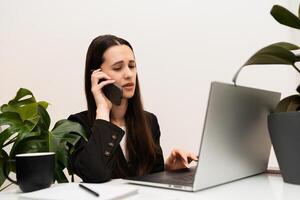 Upset woman speaking on the phone while working on computer laptop in office with white walls and green plant photo