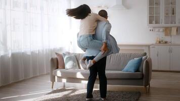 A joyful couple engages in playful interaction, with one partner giving a piggyback ride indoors. The sunlit living room features modern decor and a cozy atmosphere. video