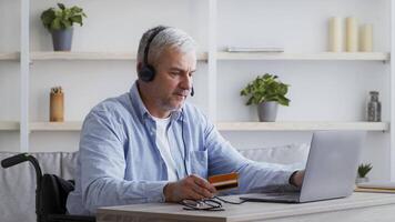 A middle aged man with grey hair is seated in a wheelchair, using a laptop at a table in his home. He wears headphones and holds a credit card, focused on online activity. video