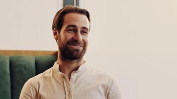 A cheerful man with a beard is sitting in a cozy cafe, smiling widely. Sunlight streams through the window, highlighting his relaxed demeanor and stylish attire. video