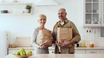 A cheerful couple stands in a modern kitchen, each holding a paper bag filled with fresh vegetables and fruits. The clean and bright space enhances their joyful moment. video