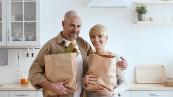 A cheerful couple stands in a bright kitchen, holding paper bags filled with colorful vegetables. They share smiles, enjoying their time together after grocery shopping. video