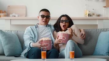 Two friends relax on a couch, wearing 3D glasses while watching a film. They share large popcorn bowls and sip orange drinks, enjoying a cozy night in together. video