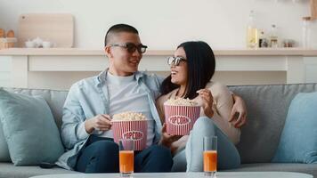 A couple relaxes on a comfortable couch, sharing popcorn and orange drinks while wearing sunglasses. They are engaged in a cozy movie night indoors, creating a fun atmosphere. video