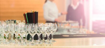 Bar counter with empty wine glasses, a container of straws, and blurred bartenders in the background. photo