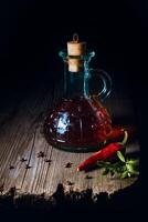 A rustic glass bottle filled with dark liquid, sealed with a cork, sits on a wooden surface next to a red chili pepper and some herbs. photo