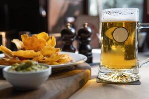 A frosty mug of beer sits next to a plate of nachos and a bowl of guacamole on a wooden board, creating a cozy and inviting dining scene, perfect for food and beverage stock photos. photo