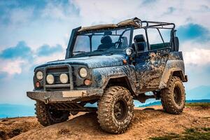 A rugged off-road vehicle covered in mud, parked on a dirt mound against a cloudy sky backdrop. photo