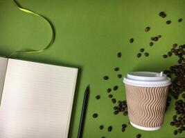 An open lined notebook with a pencil, takeaway coffee cup with a wavy pattern, and scattered coffee beans on a green background. photo