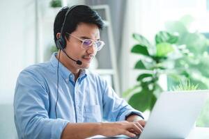 A man wearing a headset and glasses is typing on a laptop. He is focused on his work and he is in a professional setting photo