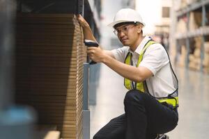 A man in a safety vest is sitting on the floor in a warehouse. He is holding a barcode scanner and looking at a box photo