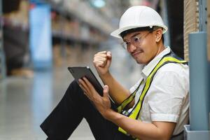 A man wearing a hard hat and safety vest is sitting on the floor with a tablet in his hand. He is smiling and he is happy photo