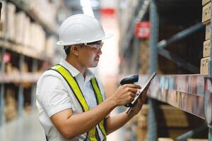 A man in a white shirt and yellow vest is using a tablet to scan a barcode. He is wearing a hard hat and safety glasses, indicating that he is working in a warehouse or industrial setting photo