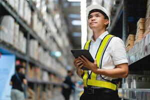 A man wearing a yellow vest and a hard hat is holding a tablet in a warehouse. He is looking up at something on the tablet photo