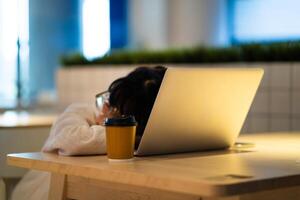A person is sleeping on a desk with a laptop and a cup of coffee. The scene suggests a sense of exhaustion or fatigue, as the person is taking a break from work or studying. The presence of the laptop photo