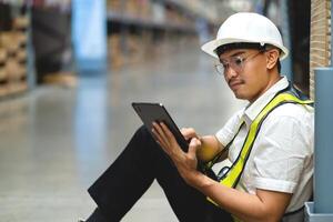 A man in a safety vest is sitting on the floor and using a tablet. He is wearing a hard hat and safety glasses photo