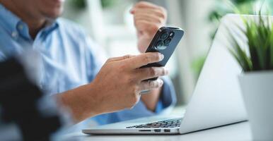 A man is using his cell phone while sitting at a desk with a laptop. The man is smiling and he is happy photo