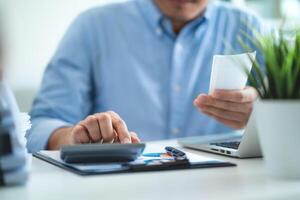 A man is sitting at a desk with a calculator and a receipt in his hand. He is focused on the receipt, possibly checking his expenses or trying to figure out how much he spent photo
