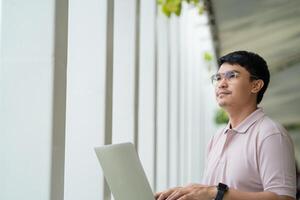 A man wearing glasses is typing on a laptop while looking out the window. He is focused on his work, but also seems to be enjoying the view outside photo