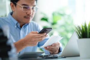 A man is sitting at a desk with a laptop and a cell phone. He is looking at the cell phone and he is checking something on it photo