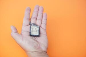 Hand holds Buddha amulet. Orange background. Concept, faith and belief of Buddhist for holy to protect from dangers, bring good luck, business prosperity and wealth. Collecting amulet as hobbies. photo