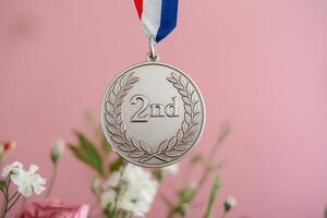 A silver medal with '2nd' inscribed, elegantly displayed against a soft pink backdrop with flowers. photo
