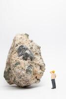 Miniature people construction worker stands next to a massive rock, scratching his head in contemplation photo