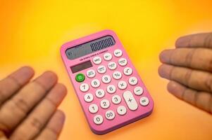 A pink calculator displaying 1000000 on its screen is held between two hands against a vibrant yellow orange background photo