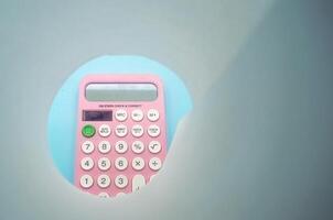A pink calculator with white buttons is viewed through a circular cutout. The calculator is placed on a light blue background, creating a unique and creative composition photo