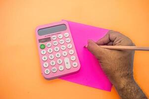 A person writes on a pink paper with a pencil next to a pink calculator on an orange background photo