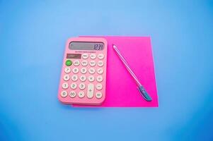 A pink calculator displaying 1234 on a blue background, accompanied by a pink pen and a pink notepad, creating a vibrant and colorful study setup photo