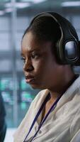 Vertical Portrait of smiling engineer hearing podcasts to make time pass faster while working in server farm to optimize it for data processing requirements. Happy woman listens songs in data center, camera A video