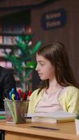 Vertical Caucasian tutor guiding elementary school girls at a library desk, providing private lessons to aid in academic development with work books and notes. Teacher discussing with scholars. Camera B. video