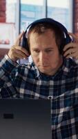 Teleworker enjoying music while researching data for company project, doing tasks in personal home office. Man sitting at desk in front of laptop and using headphones, camera B video