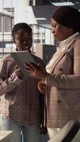 Vertical Black manager reviews company strategy with her female assistant in a high end corporate office, making key business decisions to support organizational growth. Discuss resource allocation. Camera B. video